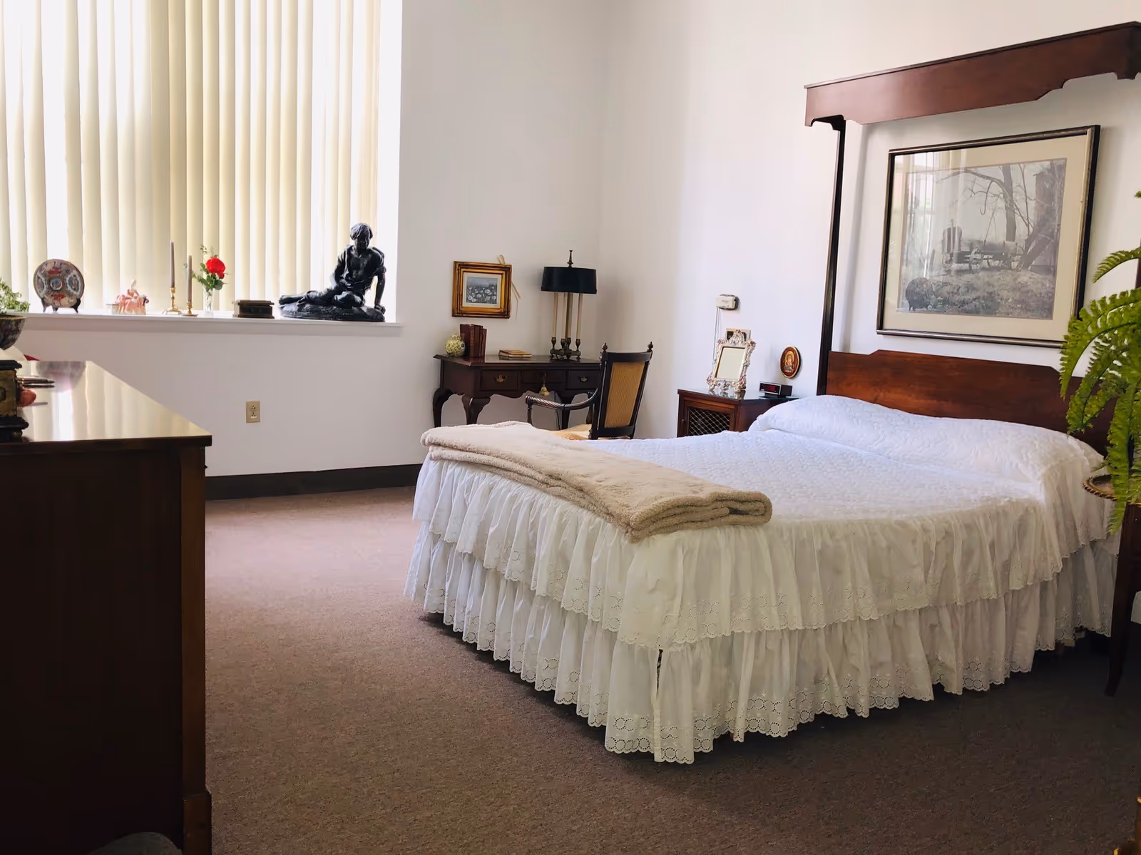 A neatly made bed with a white ruffled bedspread and a beige blanket folded at the foot. The room features a wooden headboard with a framed picture above it, a wooden dresser on the left, a small wooden desk with a chair and lamp in the corner, and a window with vertical blinds letting in natural light. Decorative items including a statue, candles, and framed pictures are placed on the window sill and furniture.
