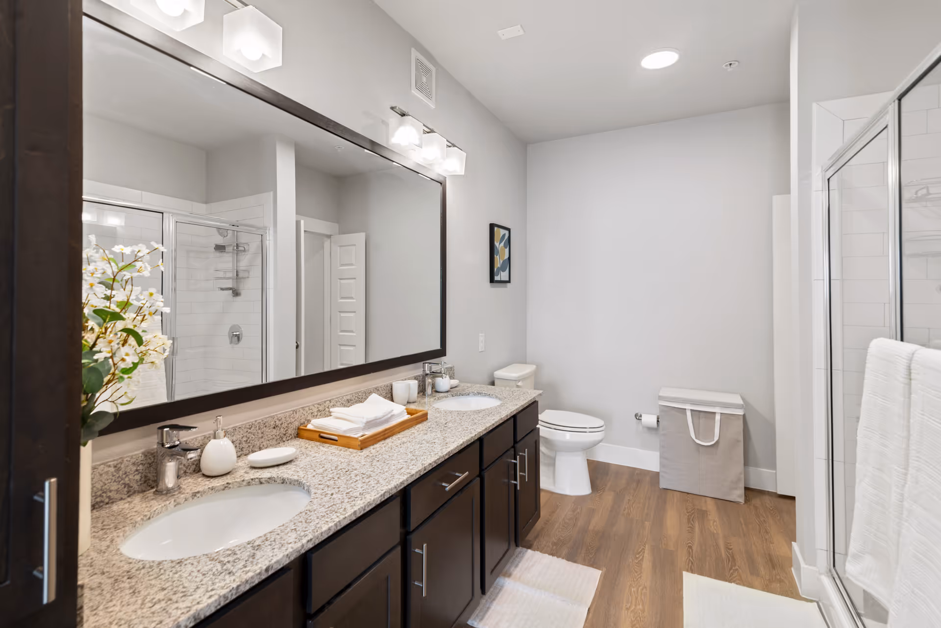 A modern bathroom with a double sink vanity featuring granite countertops and dark wood cabinets. Above the sinks is a large mirror with three light fixtures. To the right, there is a toilet next to a laundry hamper, and a glass-enclosed shower with white tiled walls. The floor is wood-style, and the walls are painted light gray. A small framed artwork hangs on the wall above the toilet.