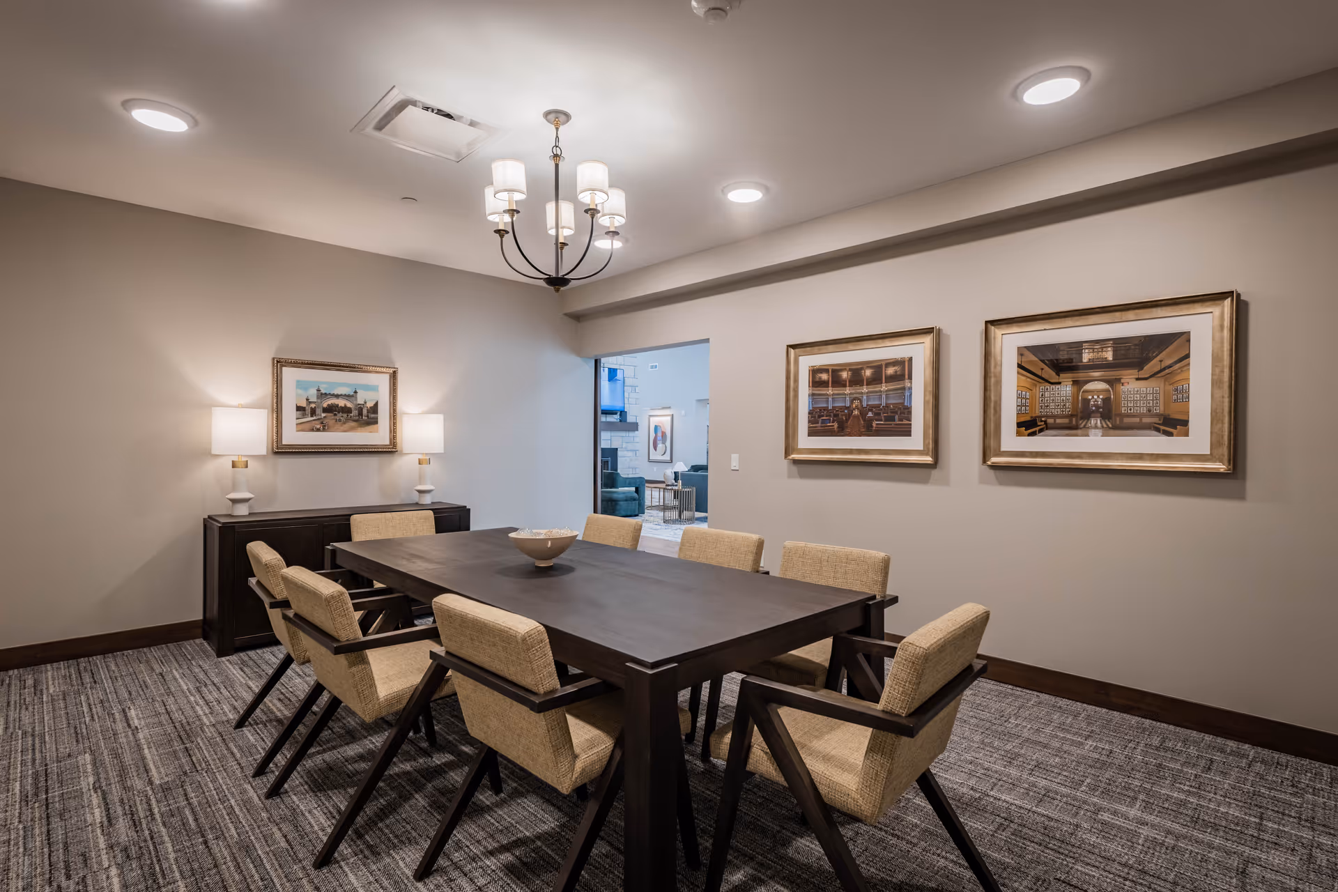 A small dining/meeting room with a long dark table surrounded by eight upholstered chairs, a chandelier overhead, framed artwork on the walls, and a sideboard with lamps.