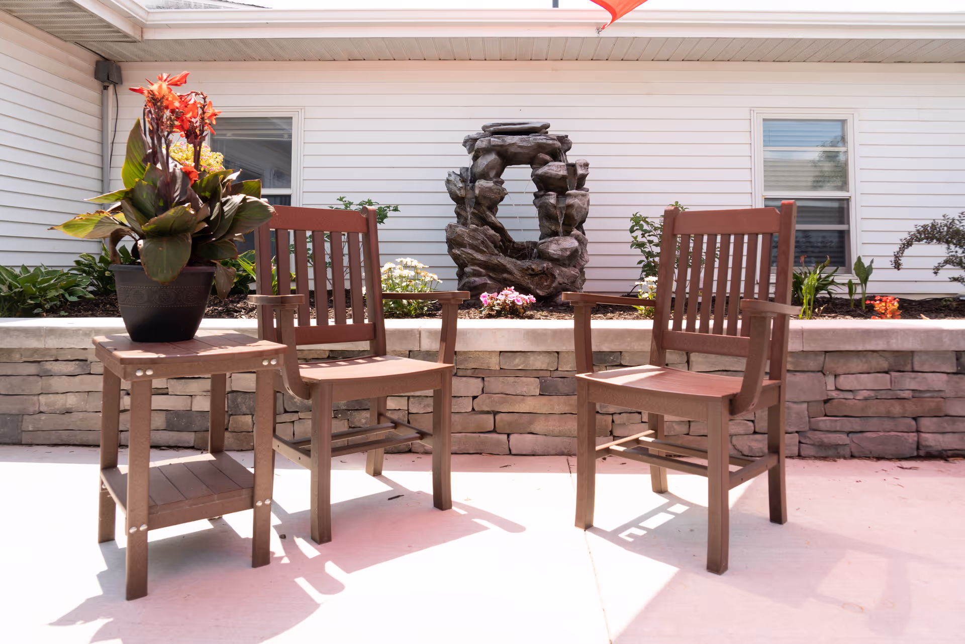Outdoor patio area with two brown wooden chairs and a small matching table holding a potted plant with red flowers. Behind the seating area is a stone retaining wall with various plants and a decorative rock water feature. The background shows the white siding exterior of a building with two windows.