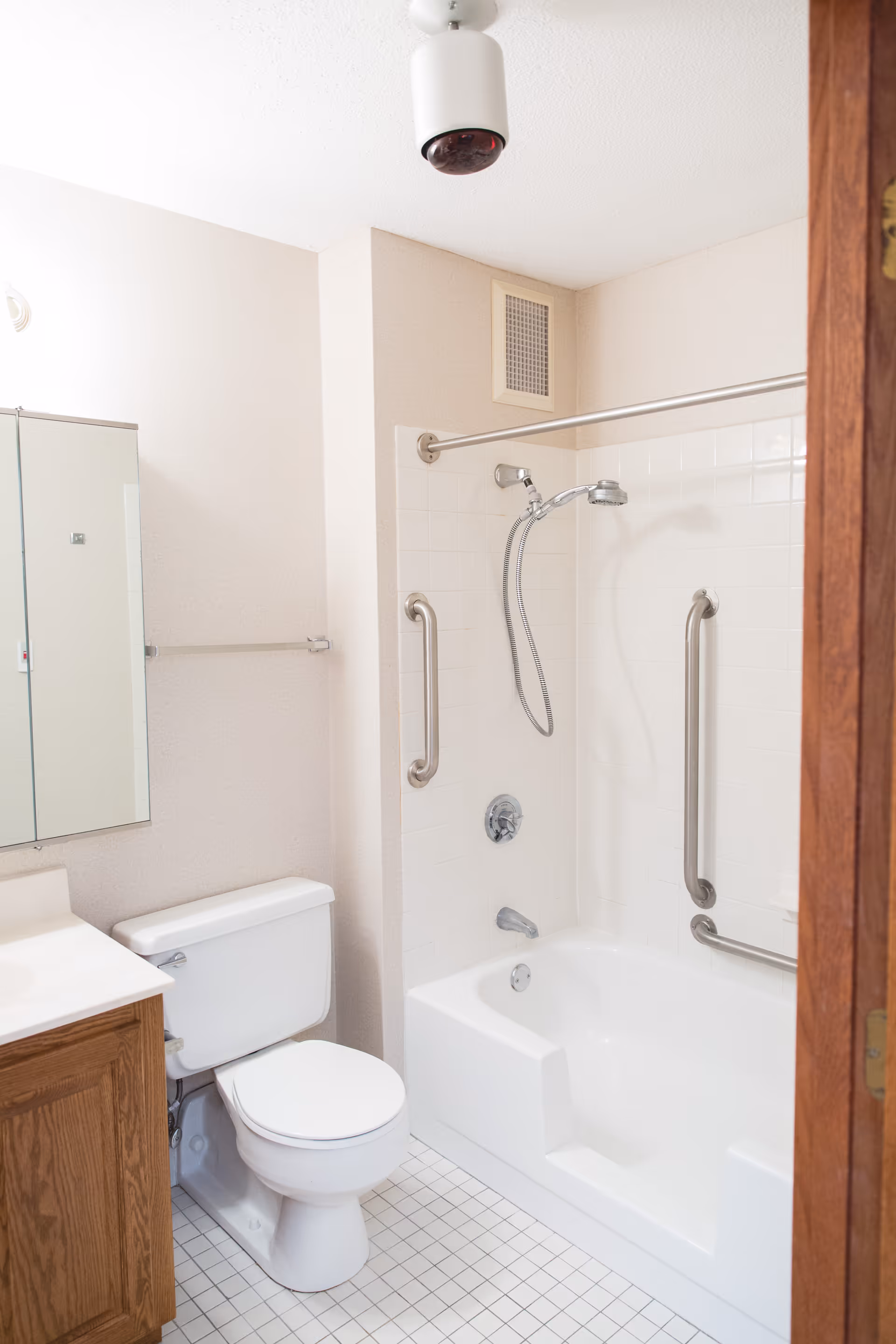 A clean bathroom with a white toilet, a wooden vanity with a white countertop, a wall-mounted mirror, and a bathtub with white tiled walls. The bathtub has two metal grab bars and a handheld showerhead. The floor is covered with small white tiles.
