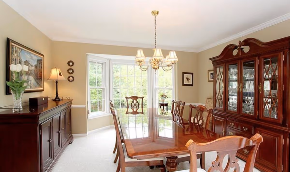 Bright formal dining room with a polished wooden table, matching chairs, a china cabinet, and a bay window.