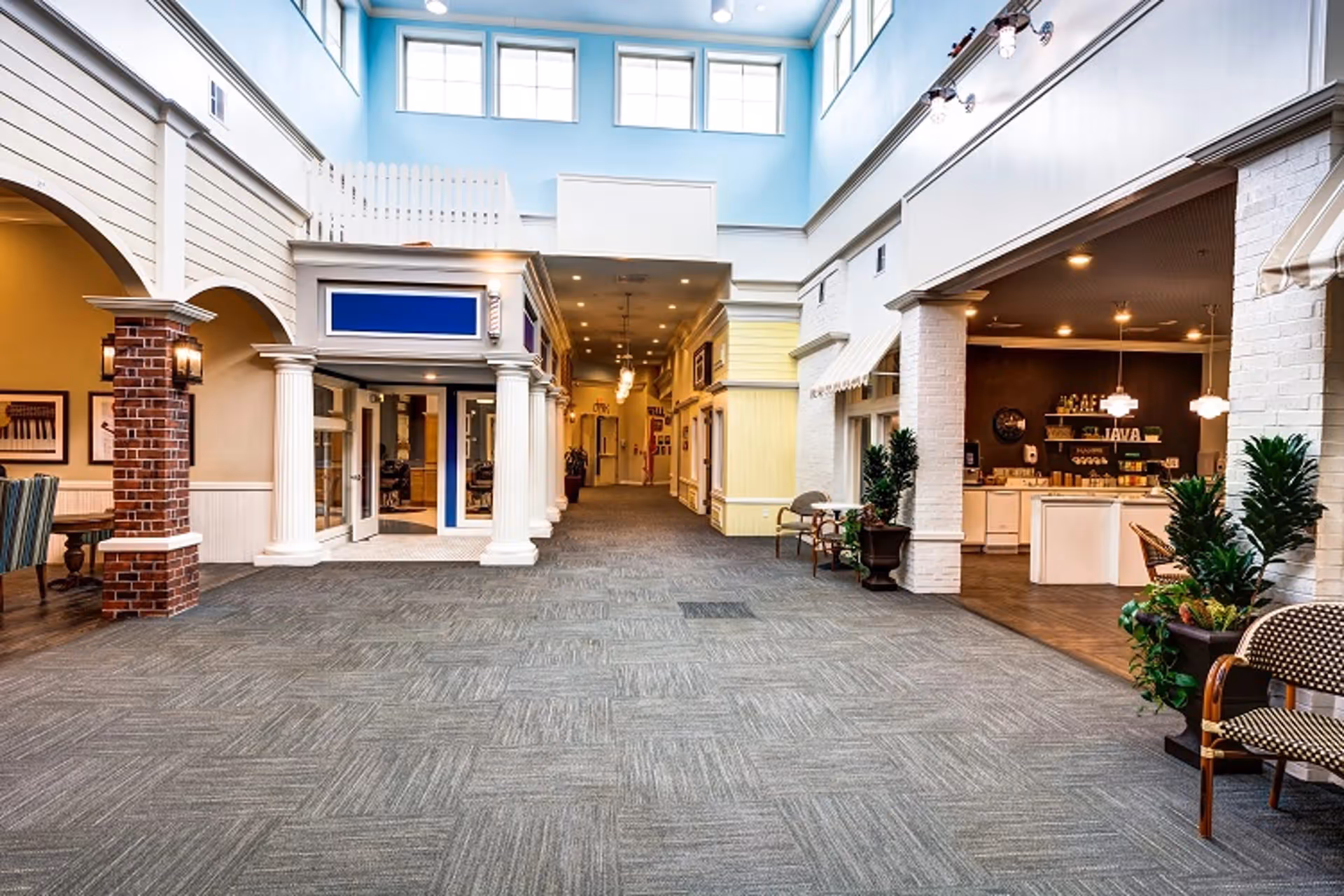 Bright two-story indoor common area with a wide carpeted hallway flanked by storefront-style entrances and a café seating area on the right.