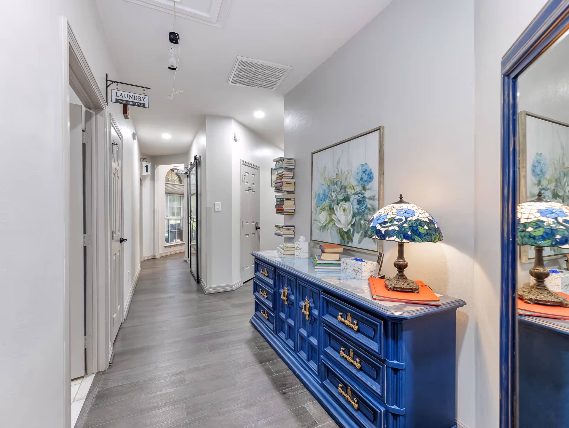 A bright hallway interior with a blue dresser topped by a stained-glass lamp and floral artwork, doors along the left wall, a stack of books, and a hanging 'Laundry' sign.