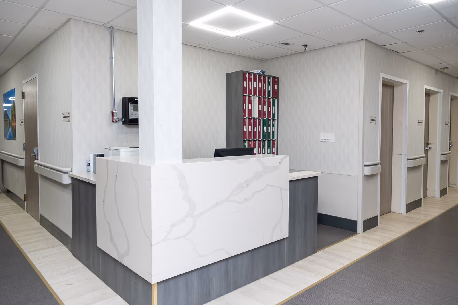 A clean and modern nursing station with a white marble-patterned counter and gray wood paneling. Behind the counter is a wall-mounted organizer with red and green folders. The area is surrounded by light-colored walls and several closed doors along the hallway.