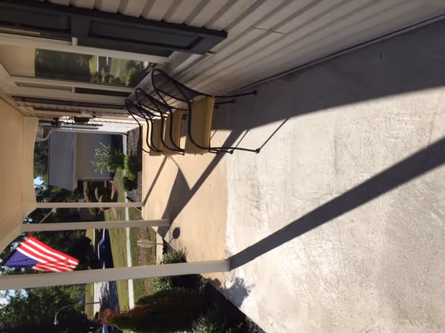 A covered outdoor patio area with two metal chairs with cushions, a concrete floor, and white siding walls. An American flag is visible in the background near a small garden and a driveway.