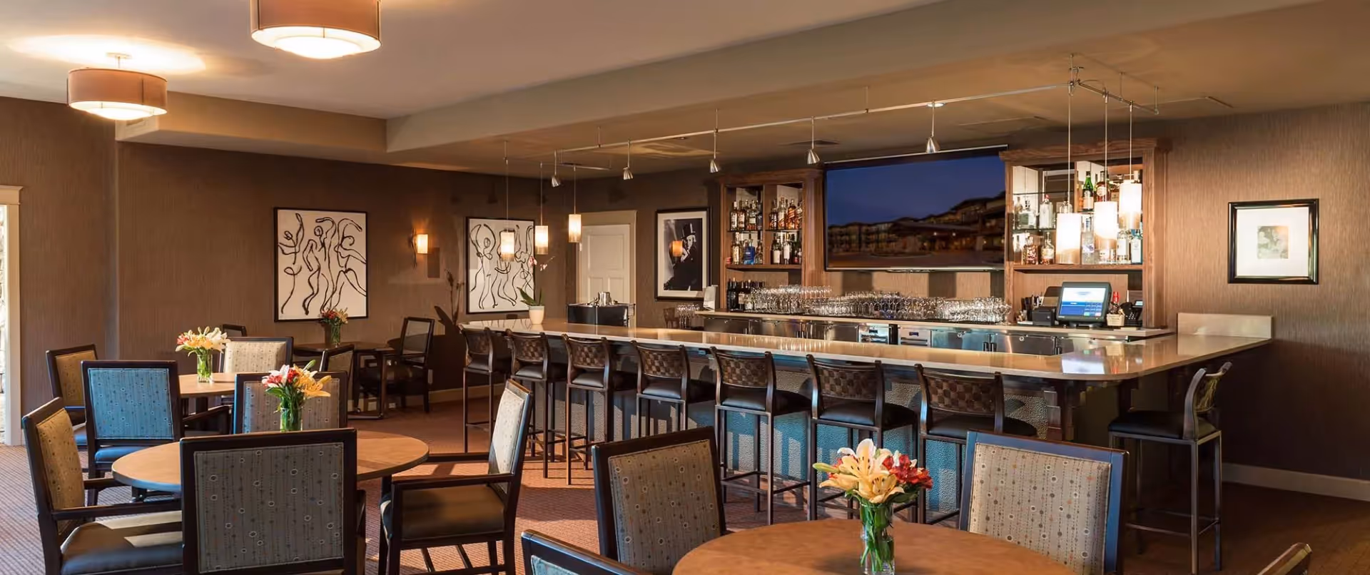 Interior view of a lounge area with a bar counter featuring high chairs, shelves stocked with bottles and glasses, a large flat-screen TV mounted on the wall, and several round tables with chairs and flower vases. The room has warm lighting and framed artwork on the walls.