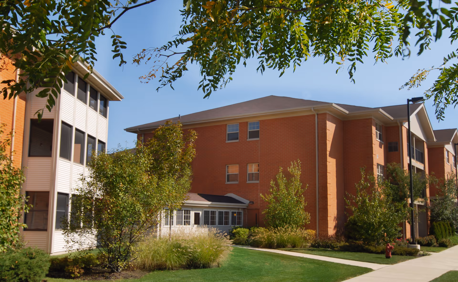 Brick multi-story senior living building with landscaped lawn, trees, and a sidewalk under a clear sky.