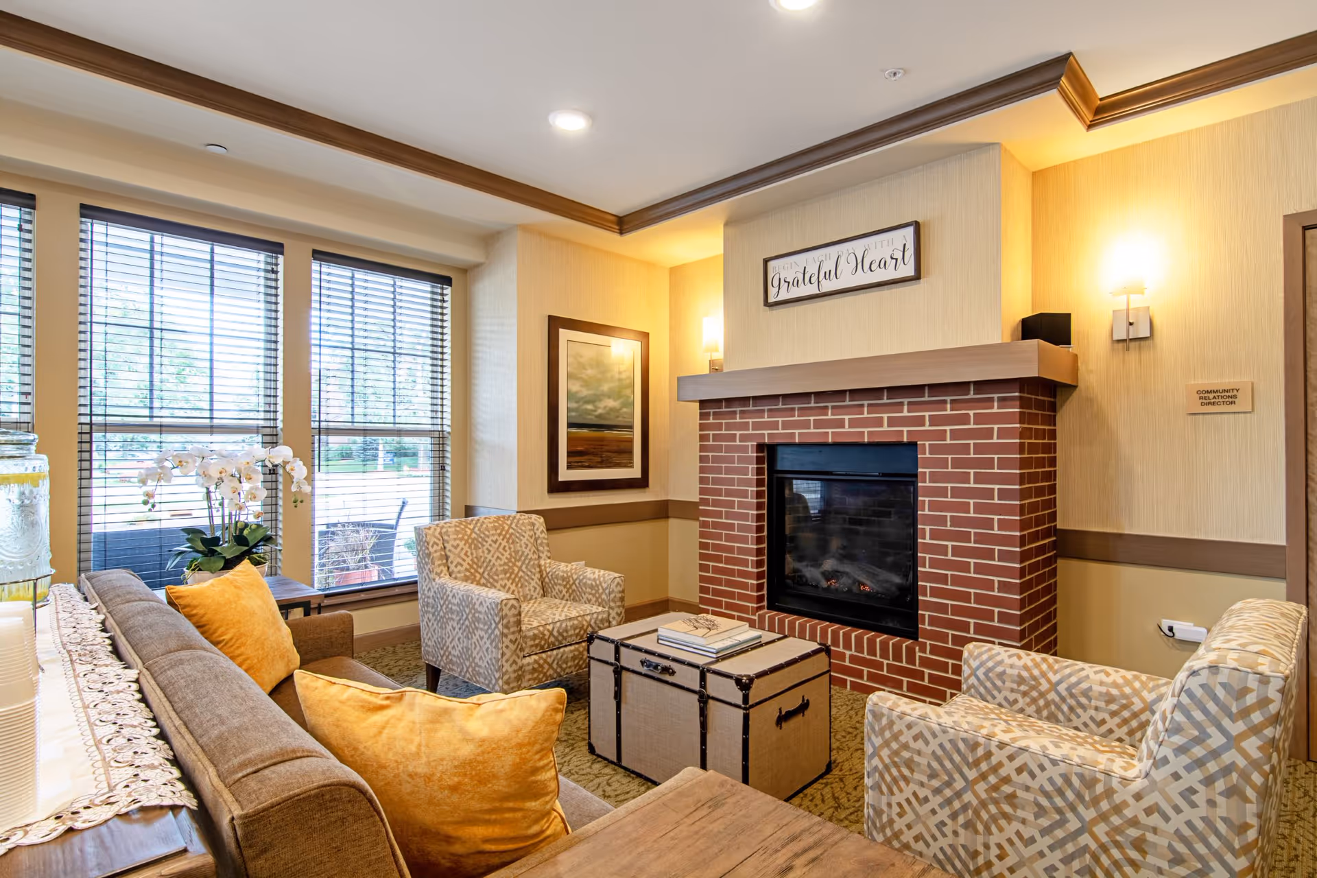 A cozy living room area with a brick fireplace, two patterned armchairs, a brown sofa with yellow cushions, a trunk-style coffee table with books on top, and large windows with blinds letting in natural light. The walls are beige with wooden trim, and there is a framed picture and a sign above the fireplace that reads 'Begin each day with a grateful heart.'