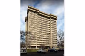 Tall beige residential high-rise building with leafless trees and parked cars in front.