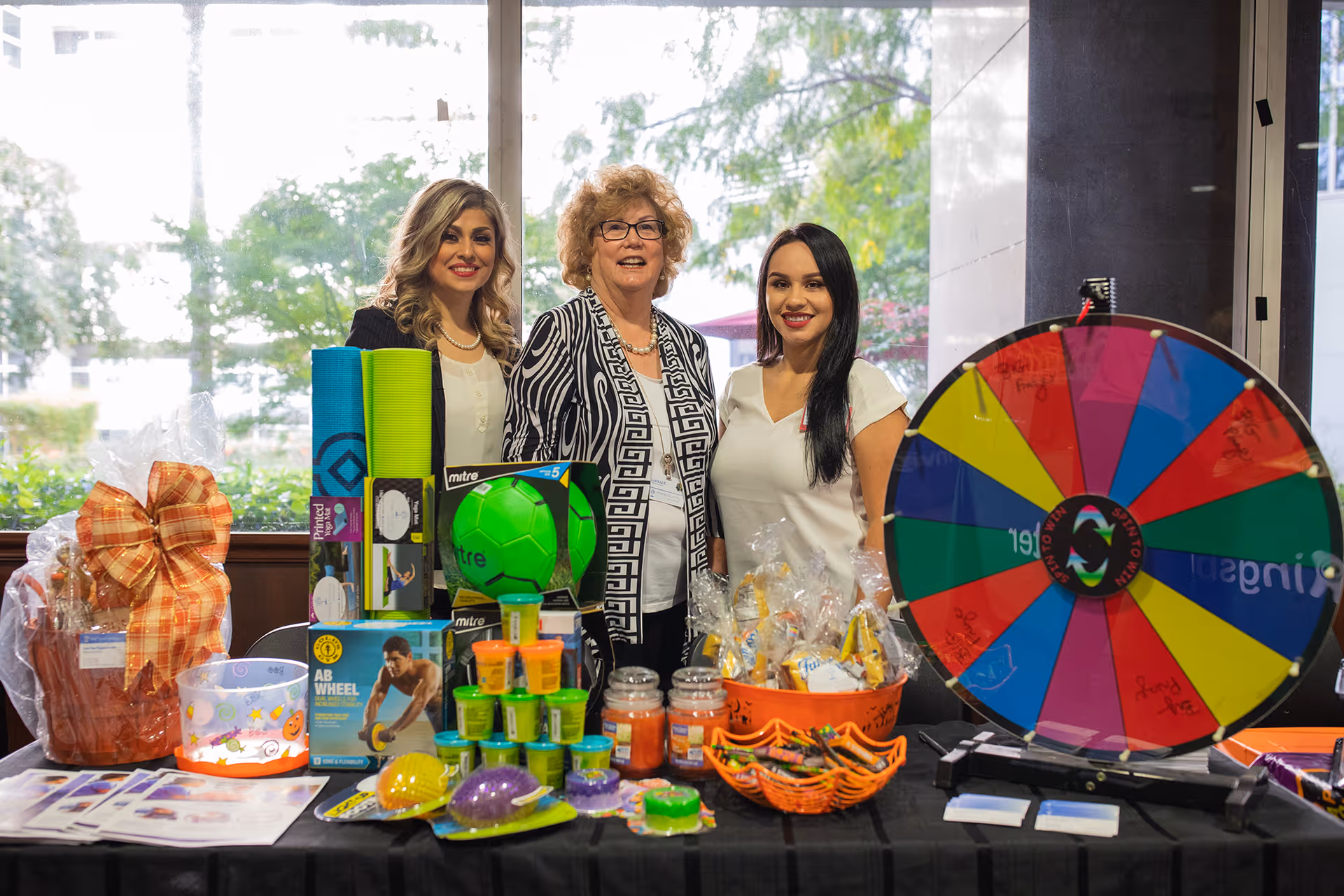 Three women standing behind a table filled with various colorful items including yoga mats, a green soccer ball, jars, small containers, baskets with wrapped treats, and a large colorful prize wheel. They are indoors with large windows showing greenery outside.
