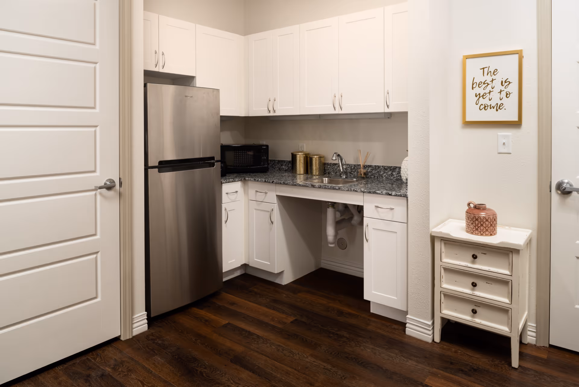 A small kitchen area with white cabinets, a stainless steel refrigerator, a microwave on the countertop, and a sink. There is a decorative sign on the wall that reads 'The best is yet to come.' A small white three-drawer cabinet with a decorative jar on top is positioned against the wall. The floor is dark wood.