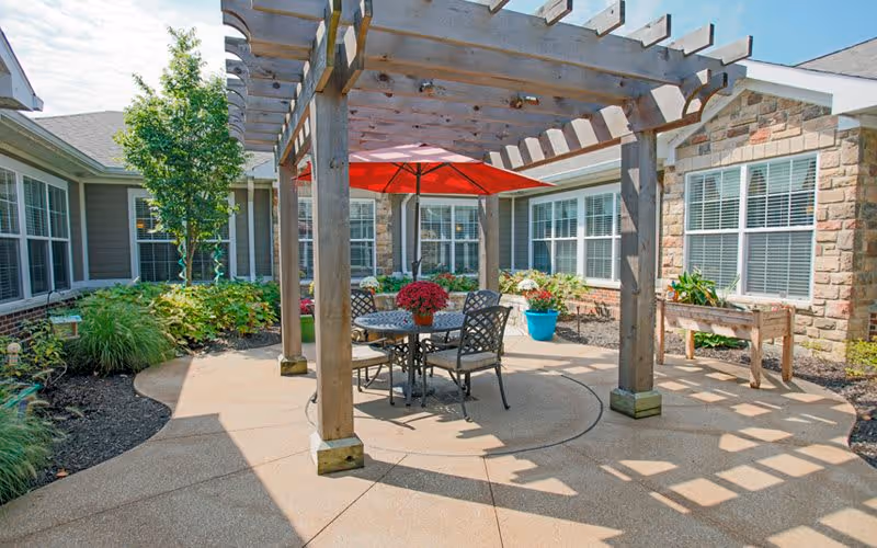 Outdoor patio area with a wooden pergola, a round metal table with four chairs, a red umbrella providing shade, and various potted plants and greenery surrounding the space. The patio is adjacent to a building with multiple windows.
