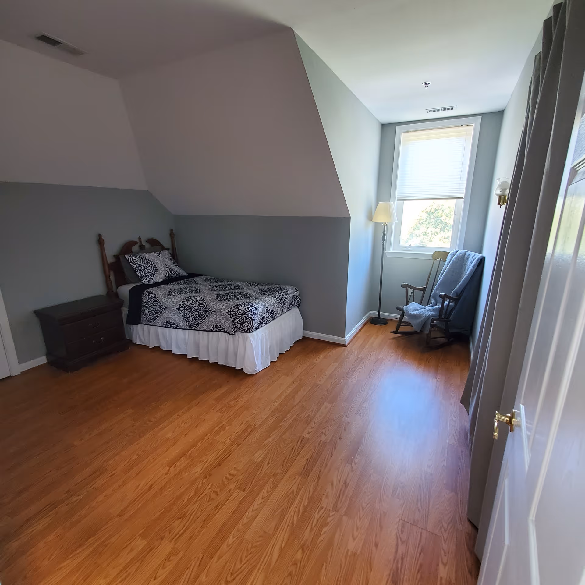 A small bedroom with light gray walls and wood flooring. The room contains a single bed with a patterned black and white bedspread, a dark wooden nightstand with drawers, a wooden rocking chair draped with a blue blanket, a floor lamp, and a window with a white blind partially drawn.