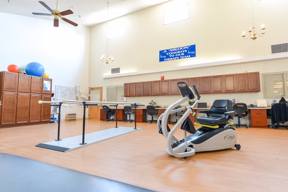 Spacious therapy and exercise room with parallel walking bars, a NuStep recumbent trainer, cabinets, desks and large exercise balls.