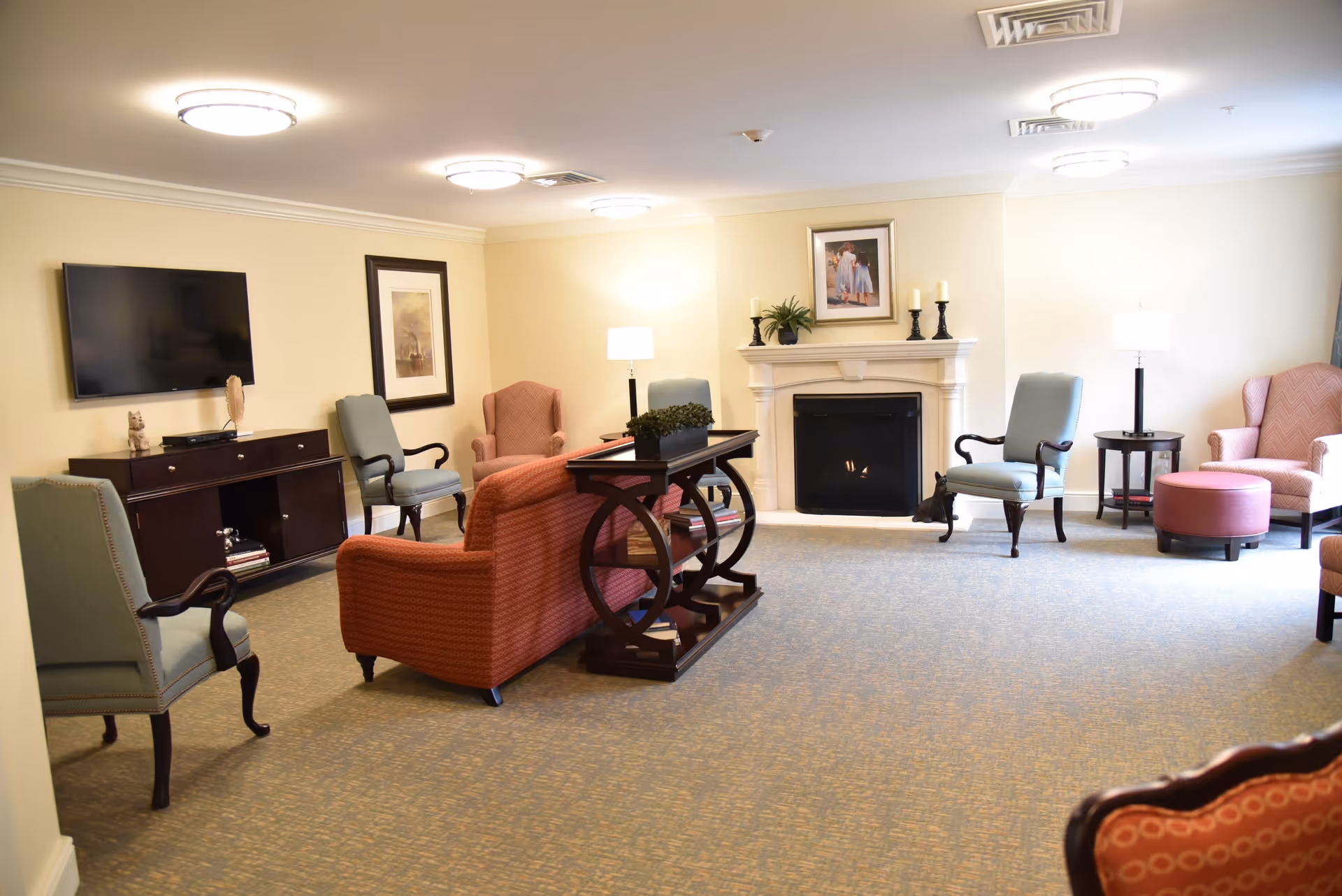 A cozy senior living room with a fireplace, several upholstered armchairs, a sofa, a TV mounted on the wall, a wooden console table with plants, and soft lighting from ceiling fixtures and lamps.