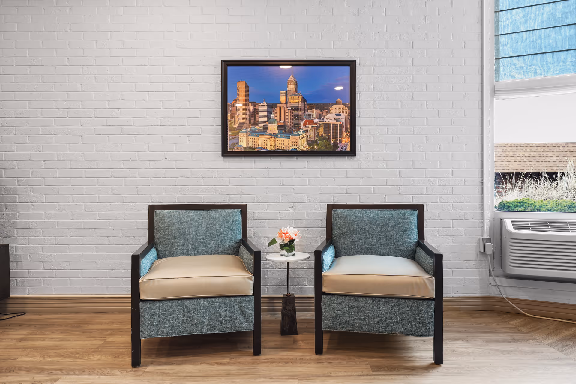 Two upholstered armchairs flank a small round table with flowers against a white brick wall beneath a framed cityscape, next to a window and air conditioner.
