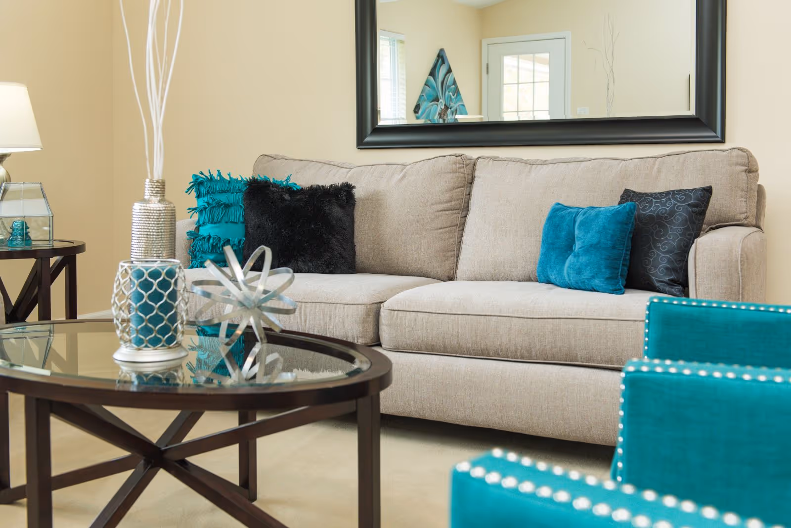 A cozy living room with a beige sofa adorned with teal and black decorative pillows. In front of the sofa is a glass coffee table with decorative items including a silver geometric sculpture and a candle holder. To the right, there are teal upholstered chairs with nailhead trim. A large mirror hangs on the wall behind the sofa, reflecting part of the room including a door and a piece of wall art.