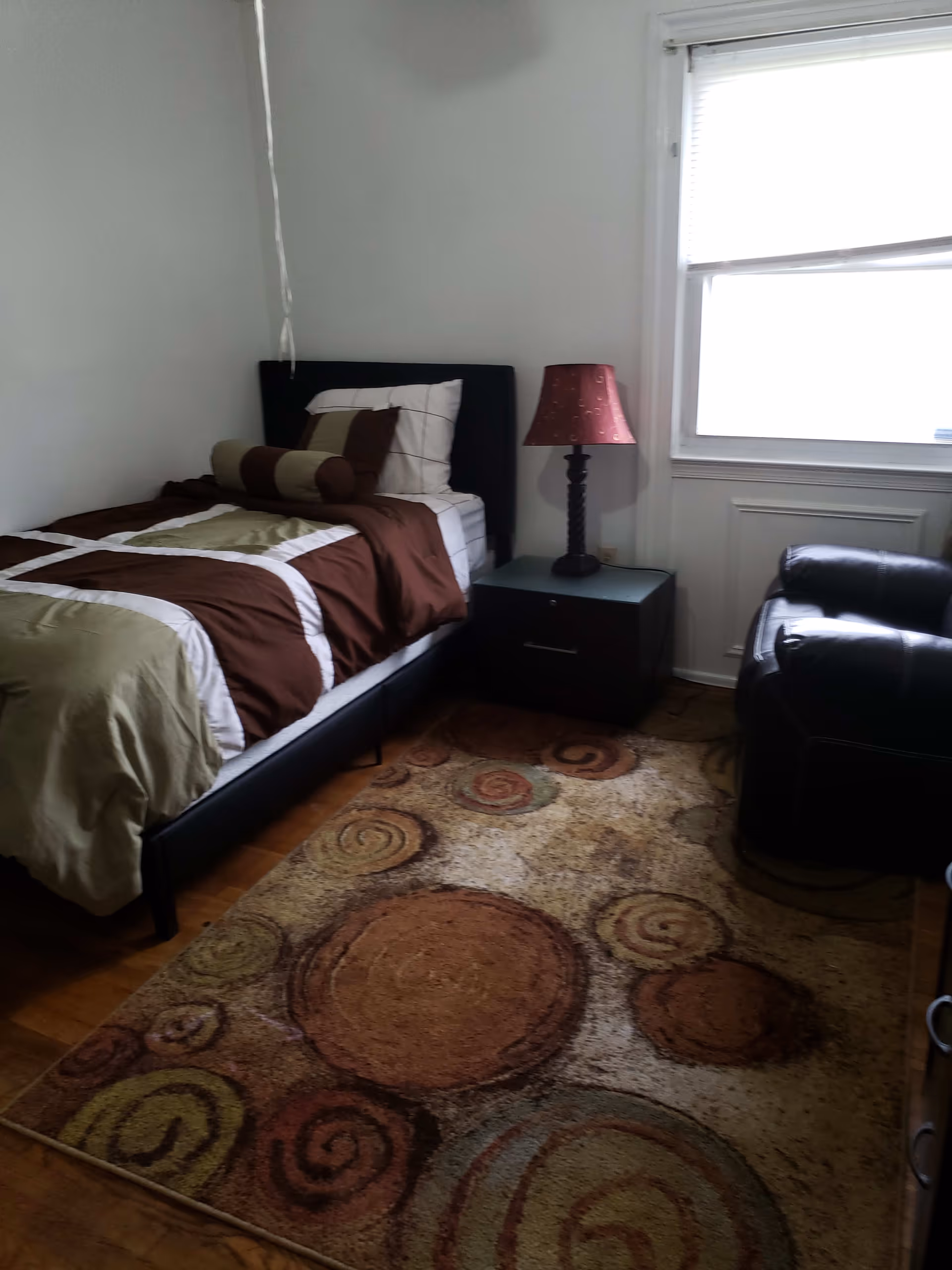 A small bedroom with a single bed made with brown, beige, and white bedding. Next to the bed is a dark nightstand with a decorative lamp on it. There is a window with blinds partially open, and a black leather armchair is positioned near the window. The floor has a rug with circular patterns in earth tones.