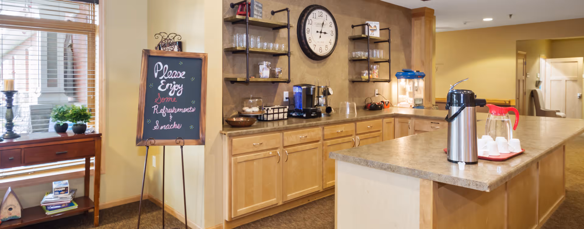 A cozy refreshment area in an assisted living facility with wooden cabinets, a countertop with coffee and snack supplies, a popcorn machine, and a chalkboard sign that reads 'Please Enjoy Some Refreshments & Snacks'. There is a window with blinds and a small table with plants and books nearby.