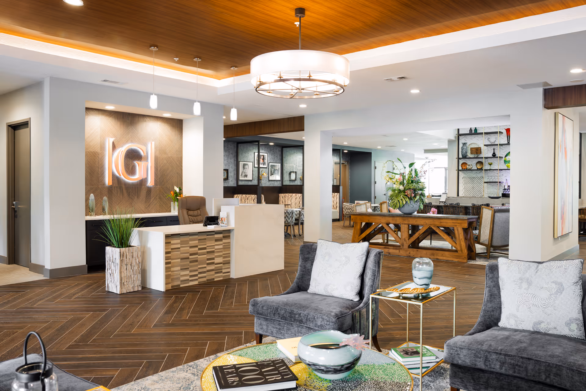 A modern and elegant senior living facility lobby with a reception desk featuring a backlit 'G' logo on a wood-paneled wall. The space includes comfortable gray armchairs with patterned pillows, a round coffee table with decorative items and books, a wooden table with a large floral arrangement, and a well-lit ceiling with pendant and chandelier lighting. The background shows a seating area with framed artwork and shelves with decorative objects.