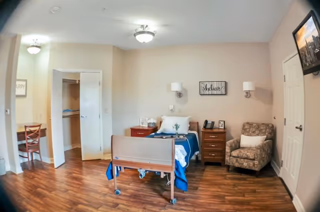 A senior living facility bedroom with a single hospital-style bed covered with blue bedding, a wooden nightstand with a lamp and telephone, a patterned armchair with a white pillow, a wall-mounted TV, and an open closet with shelves. The room has wood flooring and neutral-colored walls.