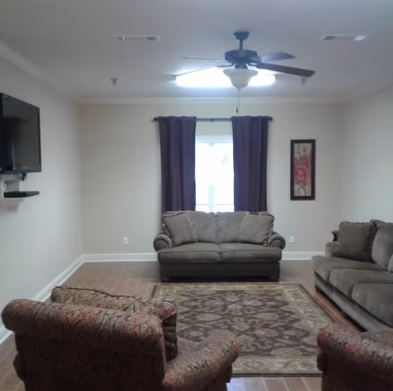 A living room with a ceiling fan and light fixture, a window with dark curtains, a wall-mounted flat screen TV, two gray sofas, two patterned armchairs, and a patterned area rug on a wooden floor.