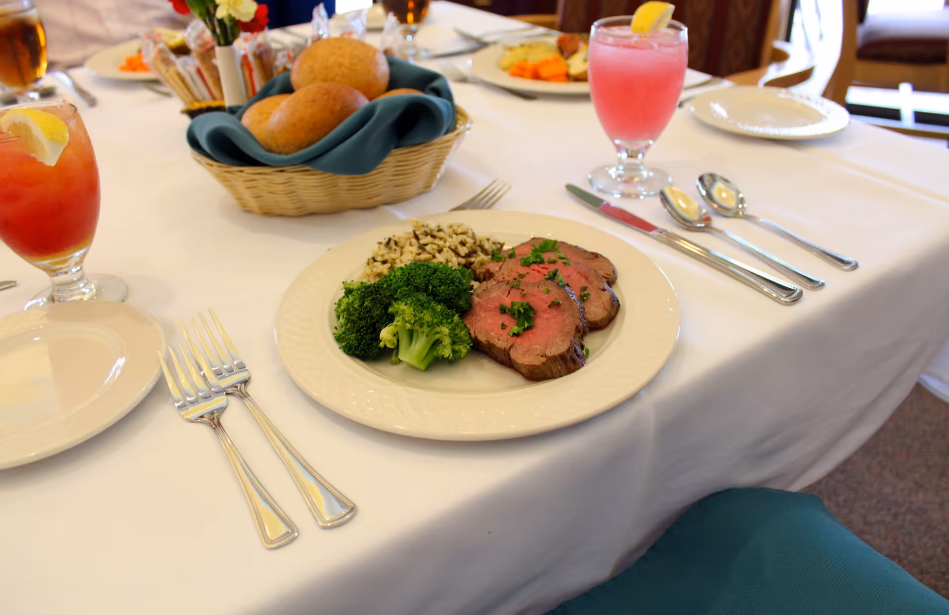 A dining table set with a white tablecloth featuring a plate with slices of roast beef, broccoli, and rice. There are two forks on the left side of the plate and a knife and two spoons on the right. A basket with bread rolls and a teal napkin is in the background, along with glasses of iced tea and a pink beverage garnished with lemon slices.