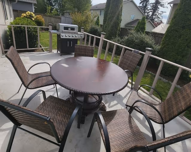 Outdoor patio area with a round wooden table surrounded by six wicker chairs. In the background, there is a gas grill and a fenced yard with green grass, bushes, and trees.