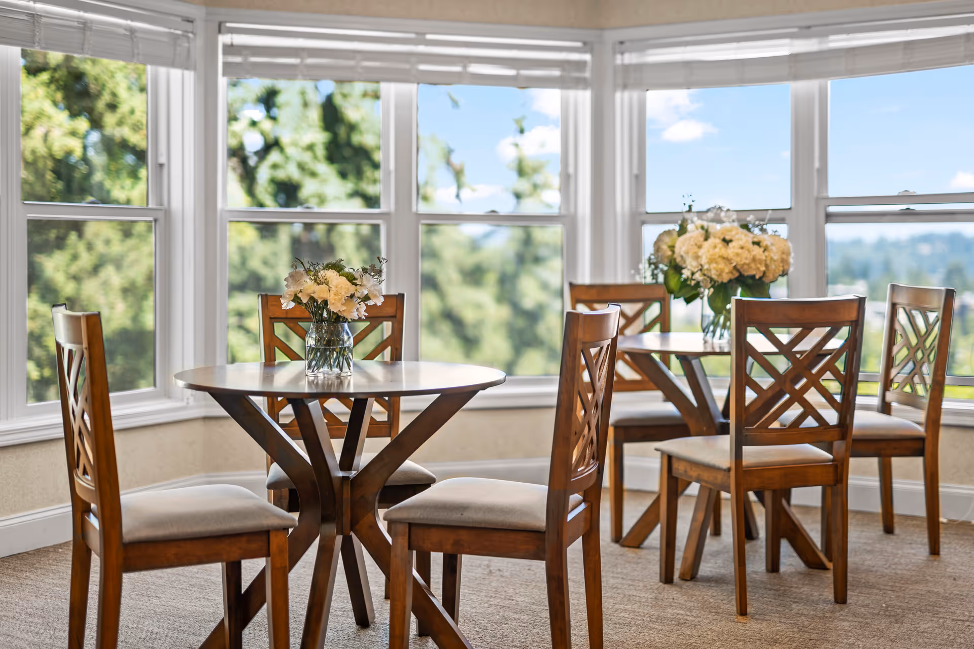 A bright dining area with two round wooden tables, each surrounded by four wooden chairs with cushioned seats. Each table has a vase with white flowers. Large windows in the background provide a view of green trees and a blue sky.