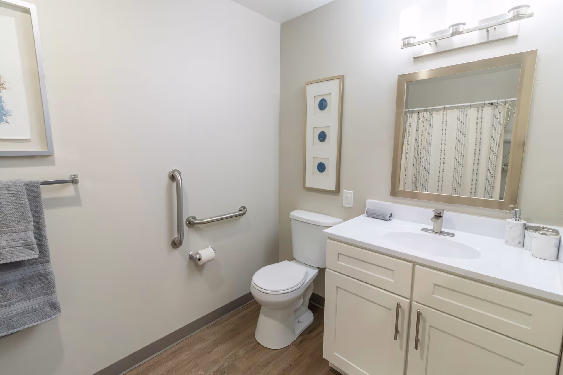 A clean and modern bathroom featuring a white toilet, a white vanity with a sink, a large mirror with a silver frame, and a light fixture above the mirror. There is a towel rack with gray towels on the left wall, a grab bar next to the toilet, and a shower curtain visible in the mirror reflection. The floor has a wood-like finish and the walls are painted light beige.