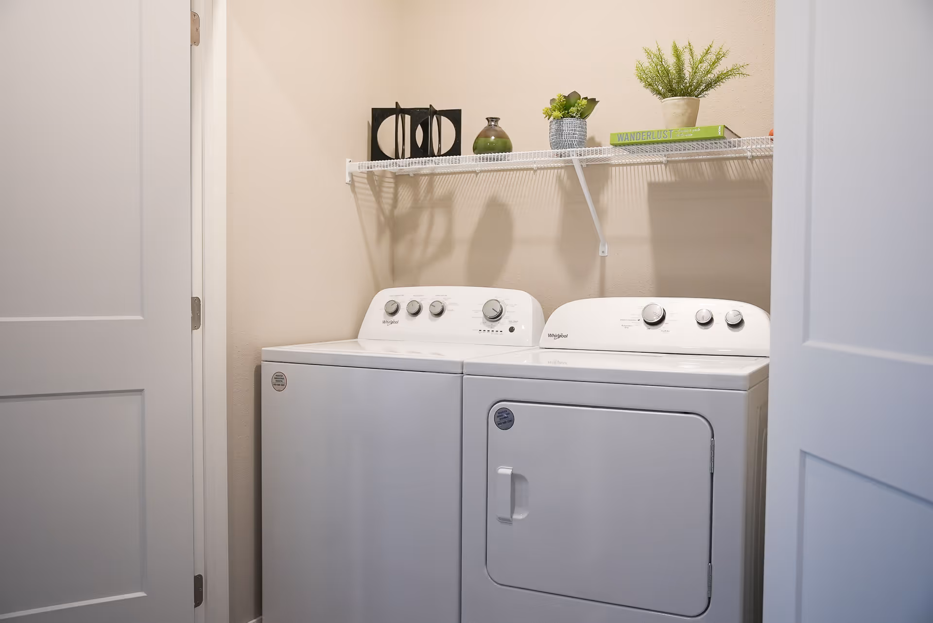 A small laundry room with a white washing machine and dryer side by side. Above them is a white wire shelf holding decorative items including a small potted plant, a green book titled 'WANDERLUST', and two small vases. The walls are beige and the room is viewed through an open door.