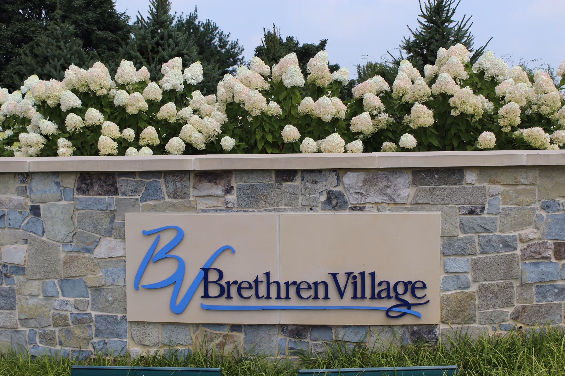 Stone wall with a sign that reads 'Brethren Village' in blue letters, with white flowering bushes and green trees in the background.