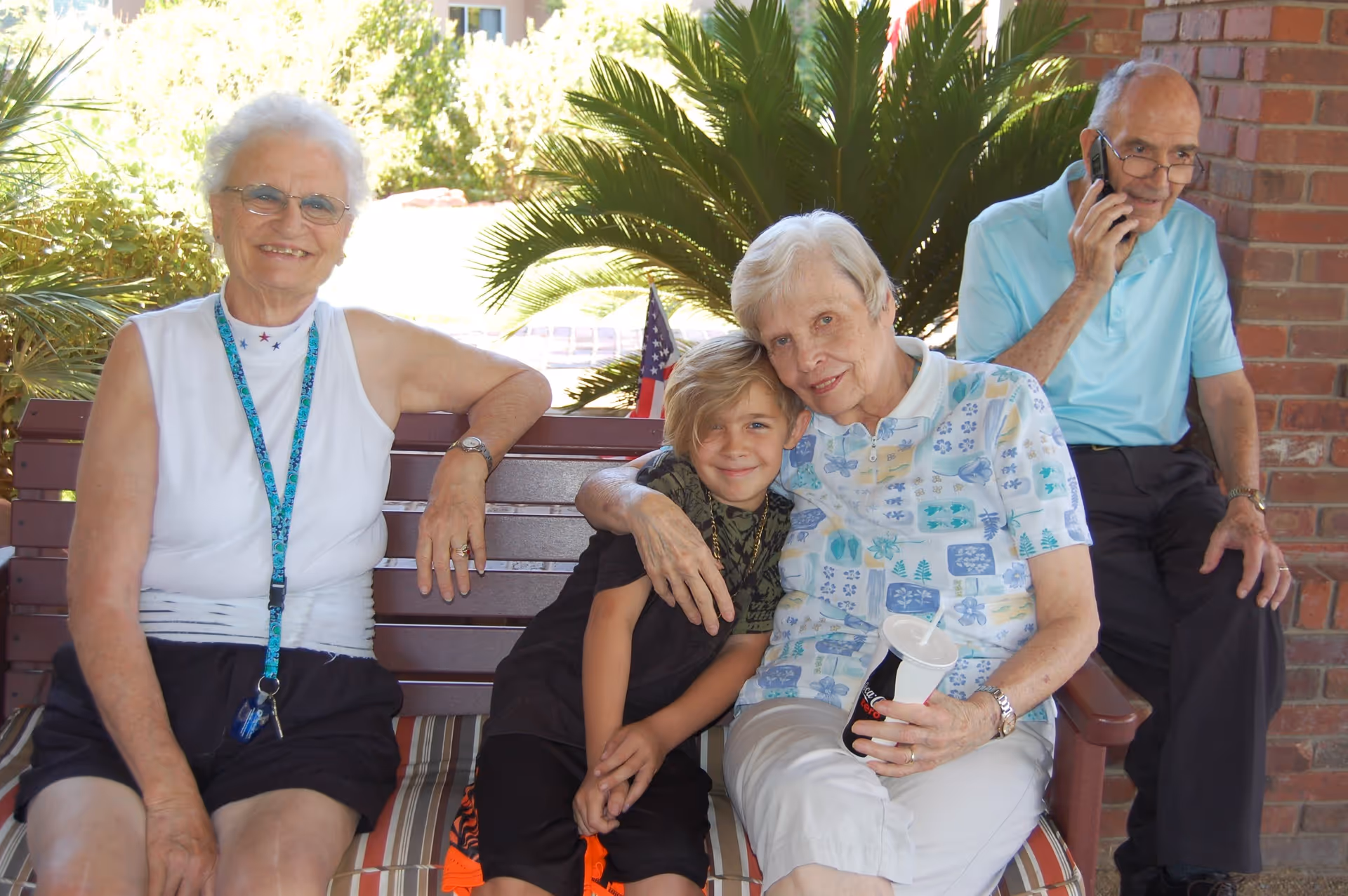 Two elderly women and a young boy sitting closely together on a bench outdoors, smiling at the camera. One elderly woman is wearing a white sleeveless top and black shorts, the other is in a patterned short-sleeve shirt holding a drink cup. An elderly man in a light blue polo shirt and dark pants is sitting nearby on the bench, talking on a cellphone. There are green plants and a brick wall in the background.