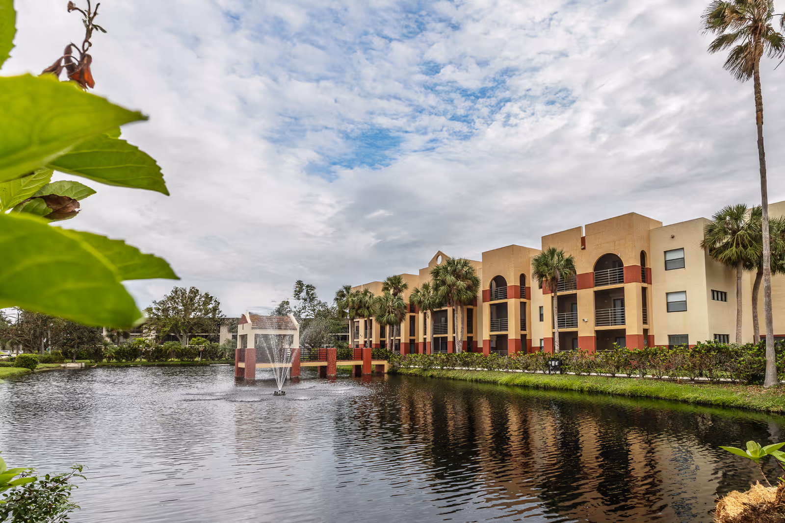A pond with a central fountain in front of a three-story beige and red assisted living building lined with palm trees under a cloudy sky.