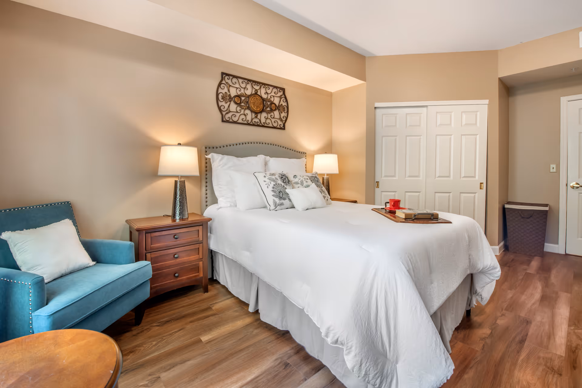 A neatly made bed with white bedding and decorative pillows in a bedroom. There are two bedside tables with lamps on each side of the bed. A blue armchair with a white pillow is positioned next to one of the bedside tables. The room has wooden flooring, beige walls, a decorative wall hanging above the bed, and a closet with white sliding doors.
