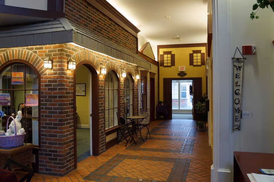 Indoor hallway with brick walls and arched windows designed to resemble a small street or storefronts. There are wall-mounted lantern-style lights, a small table with two chairs, and a basket with stuffed animals on a table. A vertical 'WELCOME' sign hangs on the right wall near the entrance to the hallway.