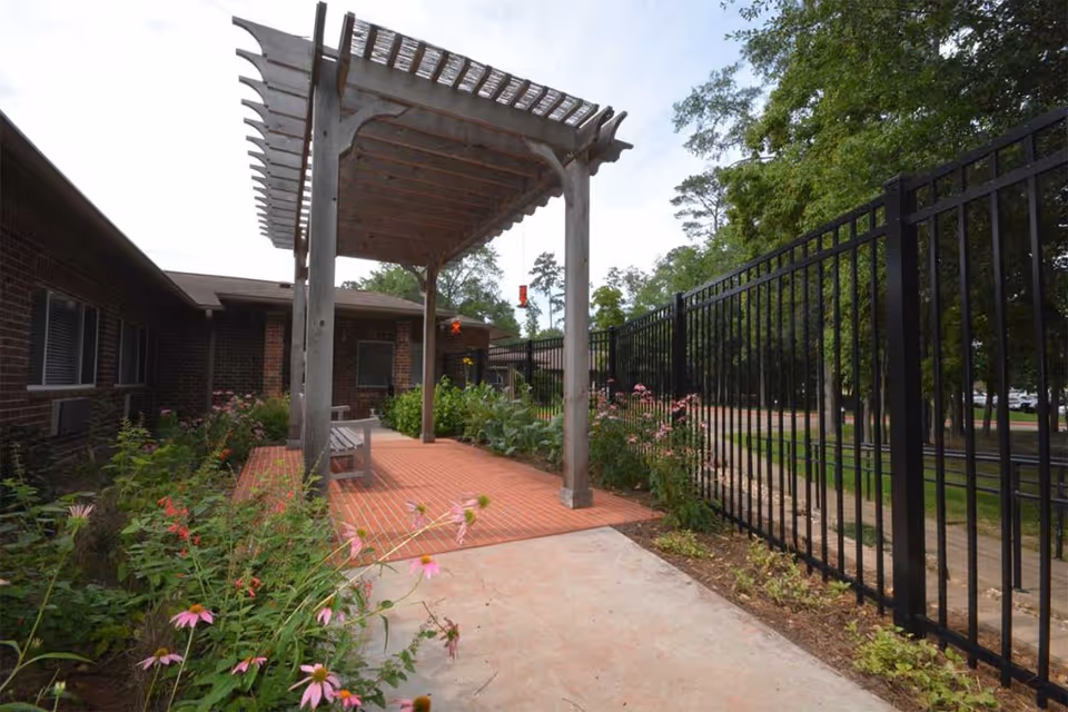 Outdoor patio area at PineCrest Retirement Community featuring a wooden pergola over a brick-paved seating area with a bench, surrounded by flowering plants and greenery, with a black metal fence on one side and a brick building on the other.