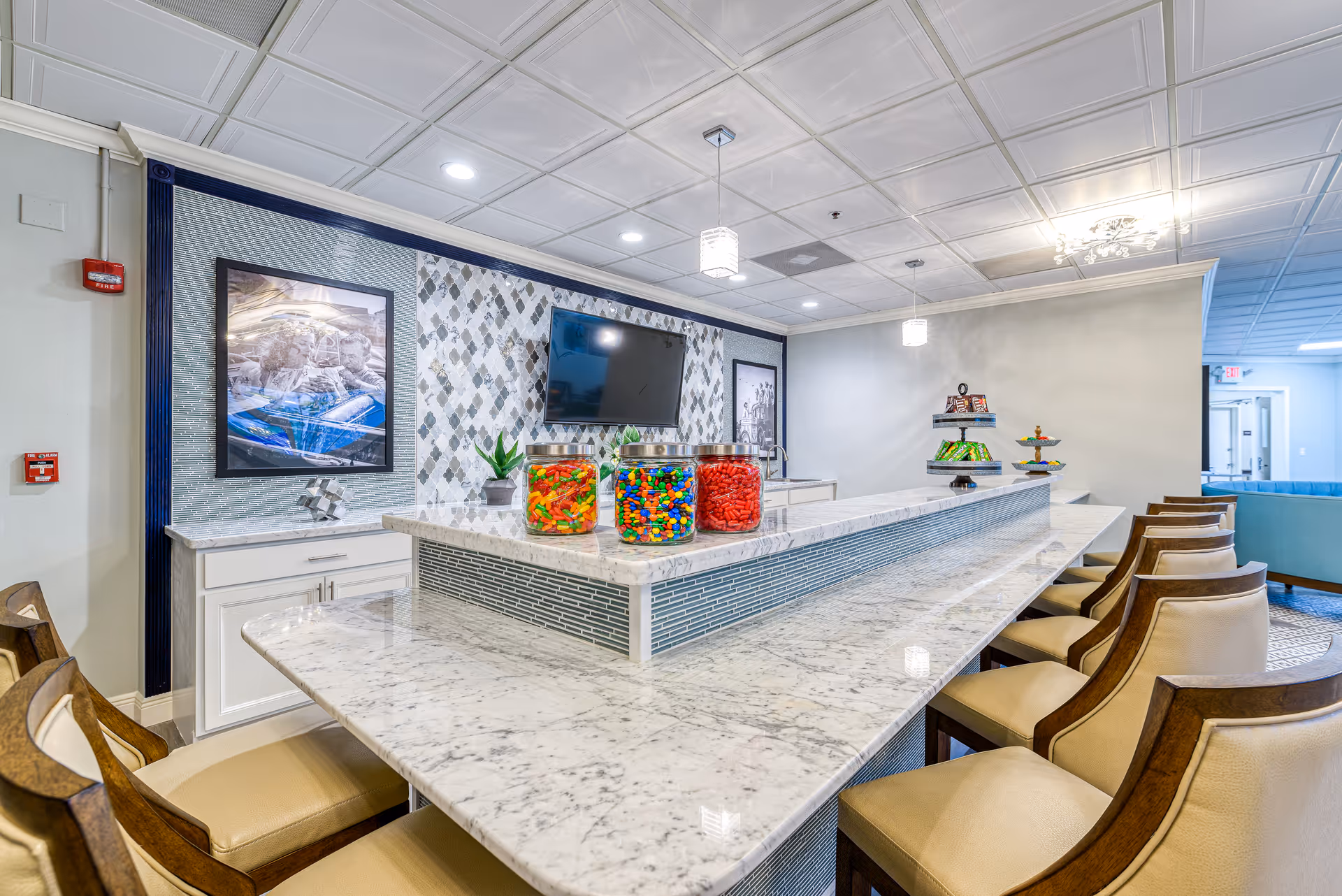 Bright communal marble bar and dining counter with stools, jars of candy, a wall-mounted TV, and seating in a senior living common area.
