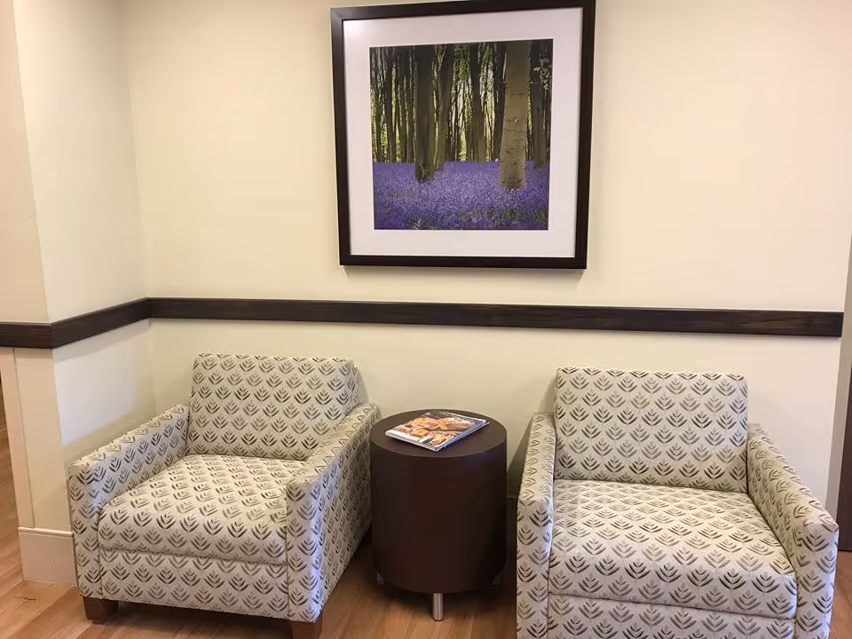 Two patterned armchairs separated by a small round dark brown table with magazines on it, placed against a beige wall with a dark wooden trim. Above the chairs hangs a framed picture of a forest with purple flowers on the ground.
