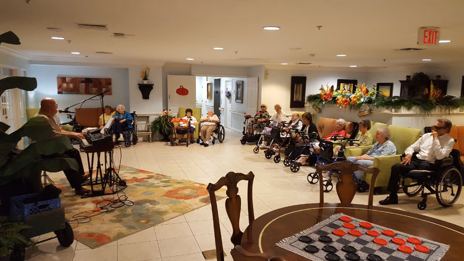 A group of elderly residents seated in a semi-circle in a common area of an assisted living facility, some in wheelchairs and others in chairs, listening to a man playing an electronic keyboard and singing. The room is decorated with plants and flowers, and there is a round table with a checkers game in the foreground.