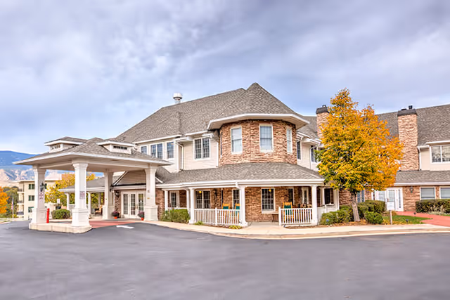 Exterior view of a senior living facility building with a covered entrance, stone and siding facade, multiple windows, and a tree with autumn-colored leaves in front.