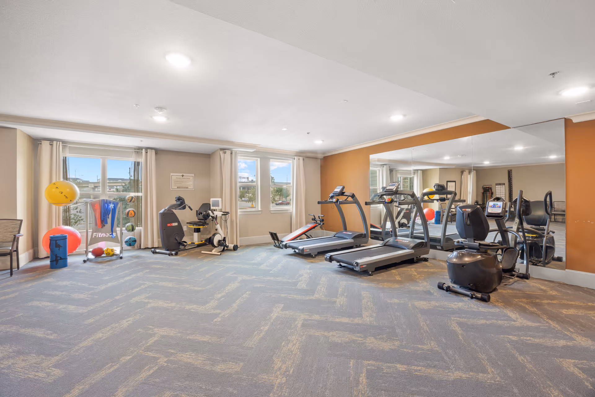 A fitness room in a senior living facility with exercise equipment including two treadmills, a recumbent bike, an arm bike, and various exercise balls and fitness accessories. The room has large windows with curtains, a large mirror on one wall, and carpeted flooring.