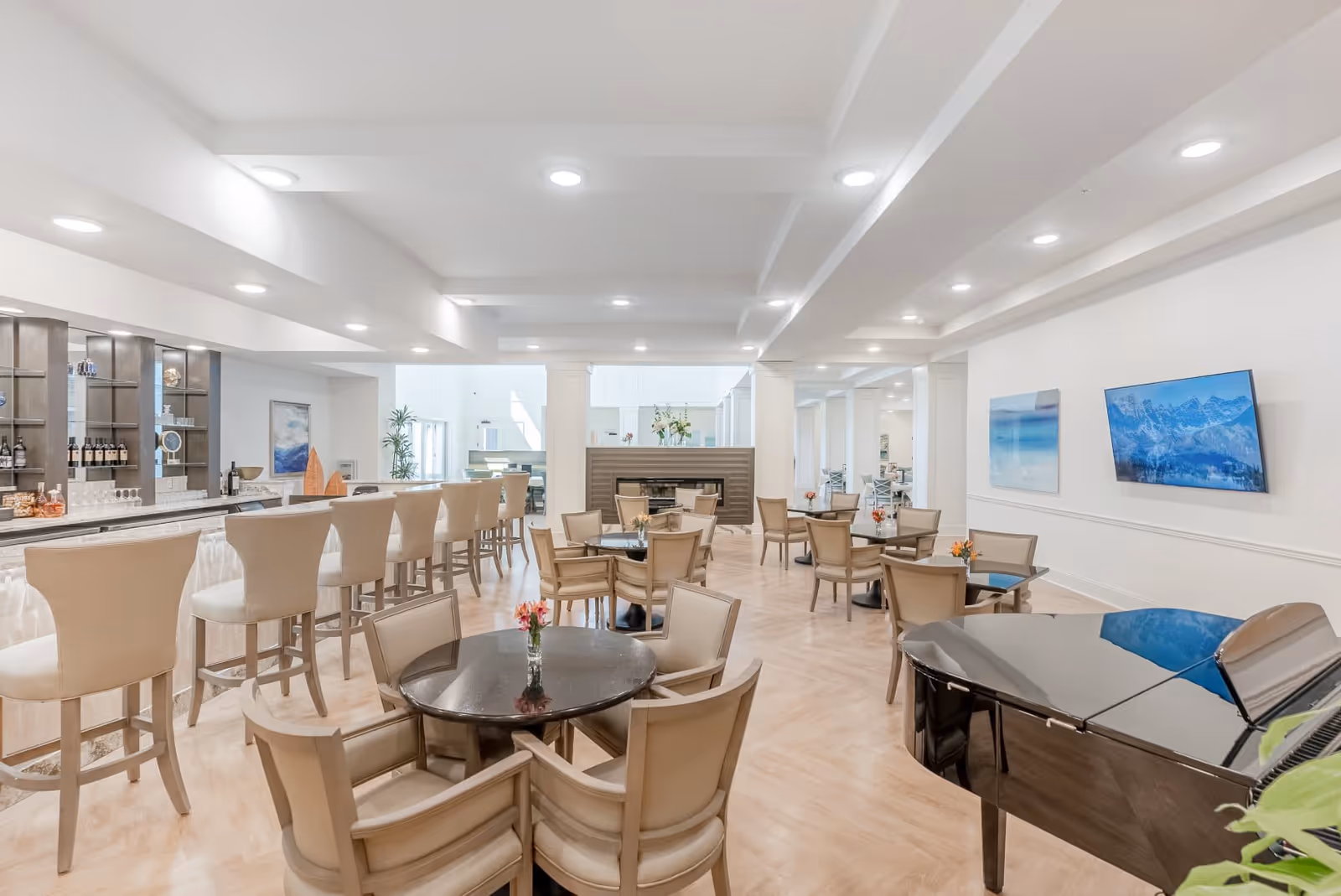 A spacious and bright common area with multiple round tables and beige chairs arranged for seating. There is a long bar counter with high chairs on the left side, a black grand piano on the right, and a fireplace in the center background. The walls are white with modern artwork and a mounted flat-screen TV. The floor is light wood, and the ceiling has recessed lighting.