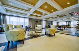 A spacious and well-lit senior living community common area with comfortable armchairs arranged around a modern coffee table. Large windows with sheer curtains allow natural light to fill the room. In the background, there are dining tables and chairs, and the ceiling features recessed lighting with a coffered design.