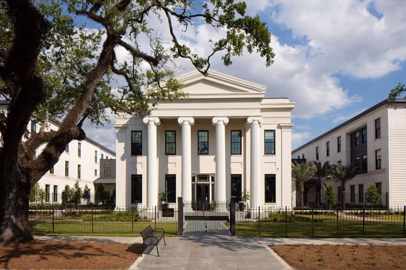 Front exterior view of The Carrollton facility featuring a grand white building with tall classical columns, large windows, and a triangular pediment. The building is flanked by two wings and surrounded by a black metal fence, with a large tree and a bench in the foreground under a partly cloudy sky.