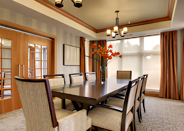 Well-lit dining room with a long wooden table surrounded by upholstered chairs, a chandelier, and large windows with curtains.