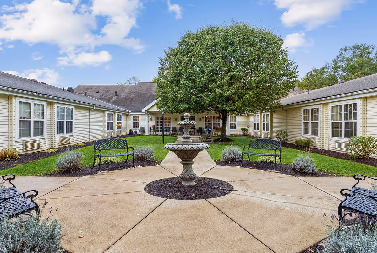 Outdoor courtyard area at The Addison of Clen-Moore Place featuring a central stone fountain, surrounded by a circular paved walkway, black metal benches, green grass, shrubs, and a large leafy tree. The courtyard is enclosed by a single-story building with beige siding and multiple windows under a partly cloudy blue sky.