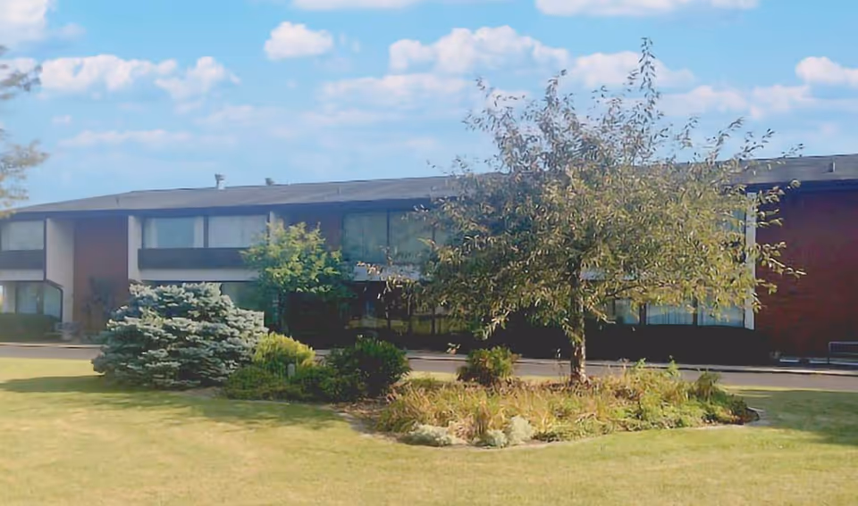 Exterior view of a two-story building with large windows, surrounded by a well-maintained lawn and landscaped garden with bushes and a tree under a partly cloudy sky.
