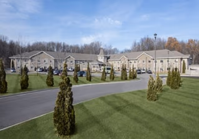 Exterior view of a large, two-story retirement community building with a curved driveway and neatly trimmed grass with small trees in the foreground under a blue sky.